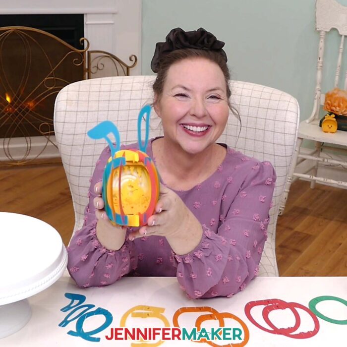 Jennifer Maker sits at her craft table, showing off a Paper Pop Up Easter Egg project, with pieces of another pop up egg on the table in front of her