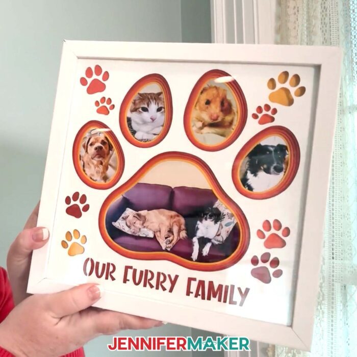 Learn to make a Photo Gallery Shadow Box with layered cardstock in JenniferMaker's tutorial! Jennifer holds up a paw print-shaped photo gallery shadow box with pictures of her pets. The box says "Our Furry Family" across the bottom.