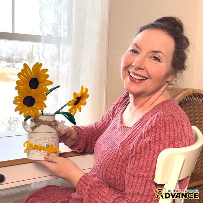 Jennifer Maker smiles as she sits with a vase of Easy Pipe Cleaner Flowers crafted with yellow pipe cleaners to look like realistic sunflowers