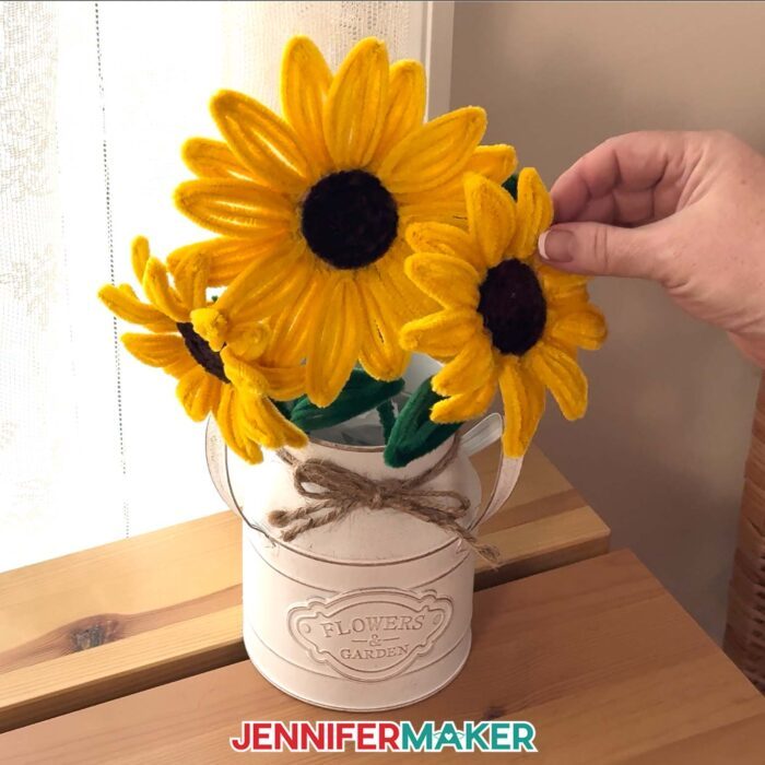 A hand touches a vase of Easy Pipe Cleaner Flowers sitting on a desk near a window. The flowers are crafted with yellow pipe cleaners to look like realistic sunflowers