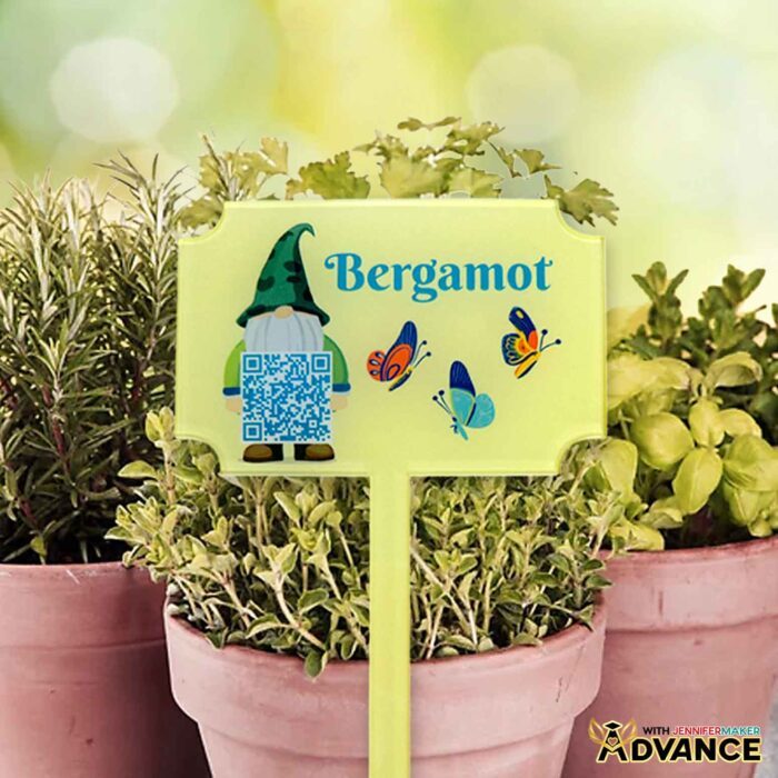 An acrylic garden marker with a gnome, three butterflies, and the word "Bergamot" sublimated on a yellow background in front of potted plants.