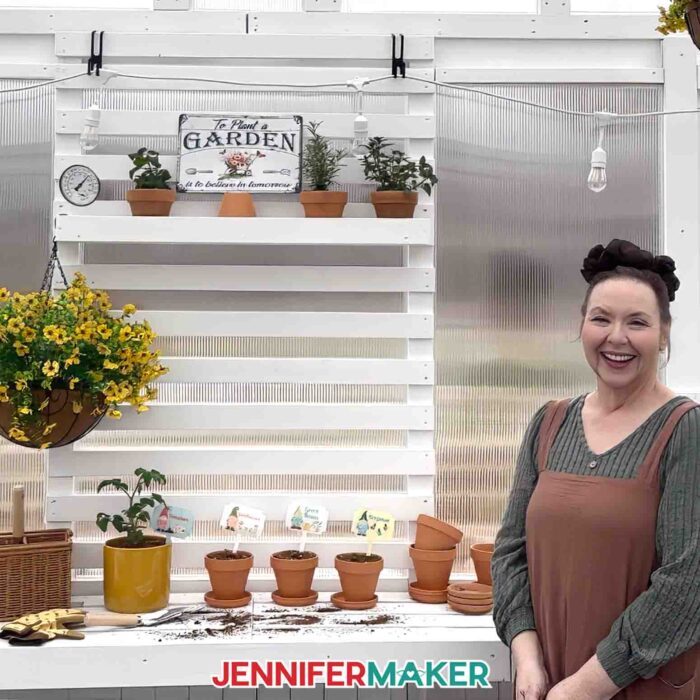 Jennifer stands in her greenhouse with pots of plants, each with a plant care garden marker placed down in the soil