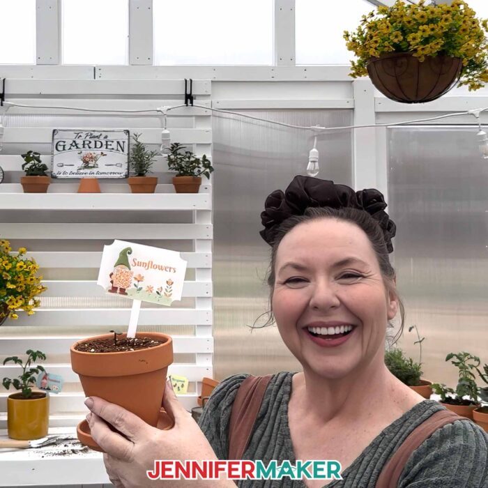Jennifer holds up a pot with soil and a garden marker labeled "Sunflowers" 