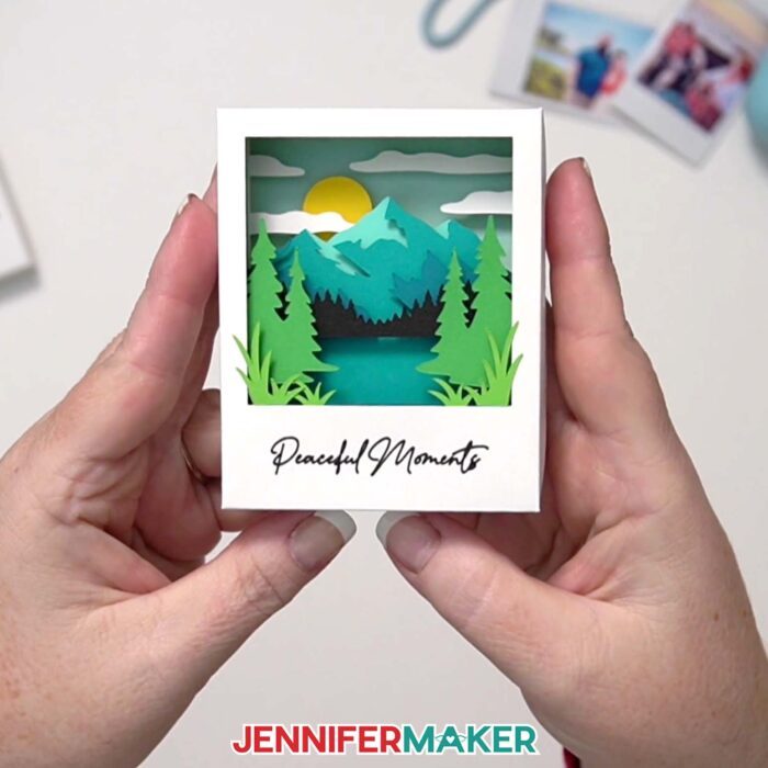 A Polaroid Shadow Box project made of colored cardstock, featuring a mountain scene and the words "Peaceful Moments" written at the bottom in Cricut pen