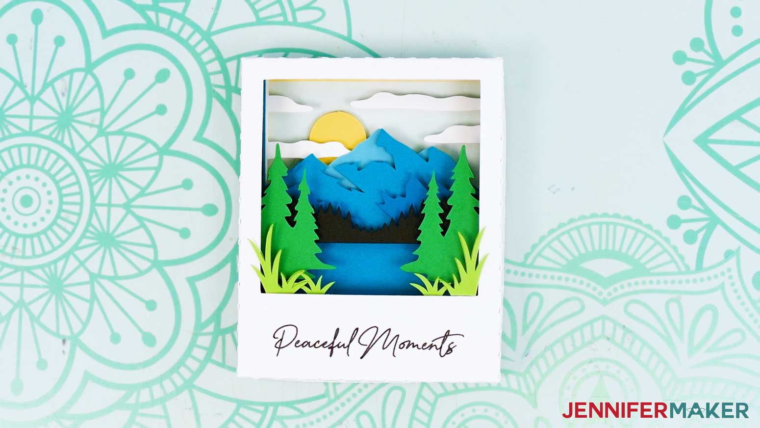 The finished cardstock Polaroid Shadow Box scene with a mountain, a sun, clouds, pine trees, a lake, and grass.