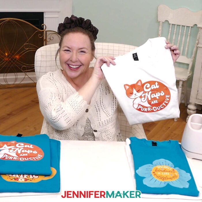 Jennifer Maker shows off a collection of turquoise and white Print Then Cut Iron-On T-shirts on display, featuring cute and cozy designs of cats, teapots, and blooms with quotes