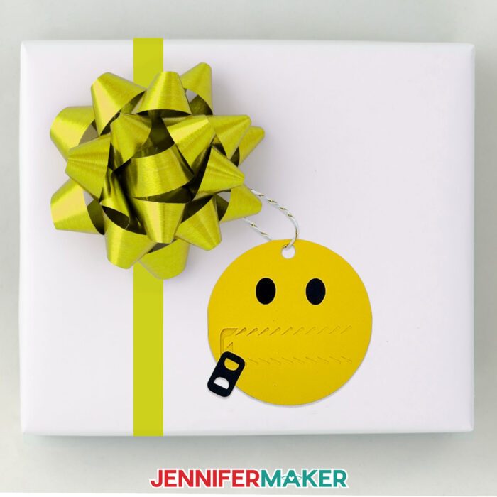 Learn how to make pull tab gift tags with JenniferMaker's tutorial! An emoji face pull tab gift tag hangs from a bow on a wrapped gift.