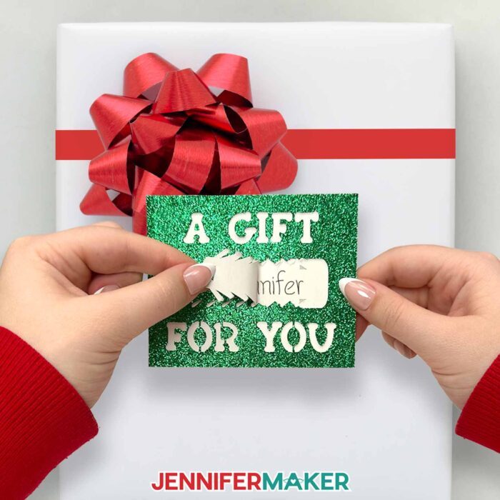 Learn how to make pull tab gift tags with JenniferMaker's tutorial! A green glitter pull tab gift tag hangs from a bow on a wrapped gift.