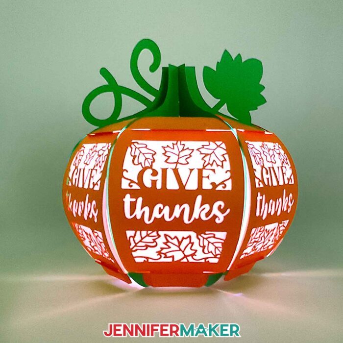 Learn how to make a 3D pumpkin lantern with JenniferMaker's tutorial! An orange paper pumpkin lantern with a green stem, vine, and leaf feature panels with intricate filigree leaf and "Give Thanks" designs cut out.
