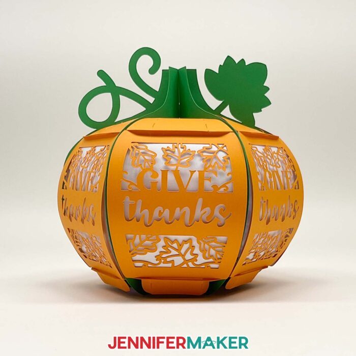 Learn how to make a 3D pumpkin lantern with JenniferMaker's tutorial! An orange paper pumpkin lantern with a green stem, vine, and leaf feature panels with intricate filigree leaf and "Give Thanks" designs cut out.