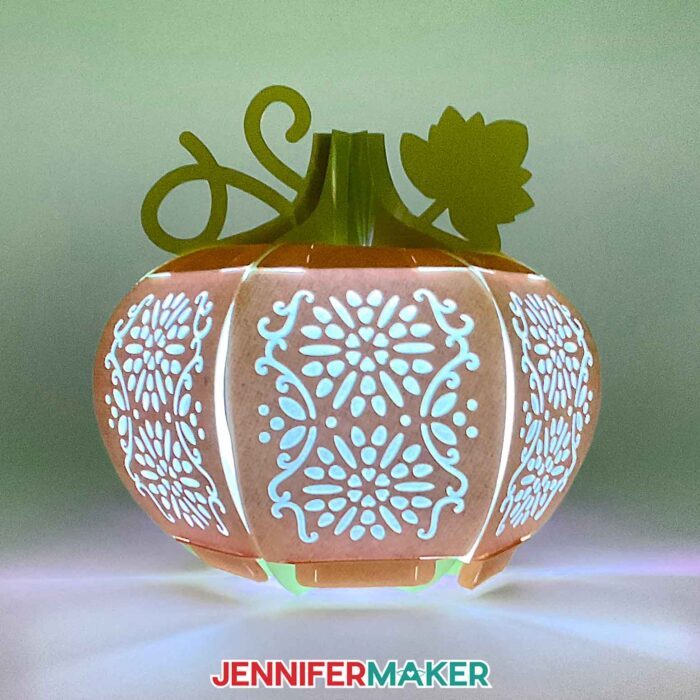 Learn how to make a 3D pumpkin lantern with JenniferMaker's tutorial! An orange paper pumpkin lantern with a green stem, vine, and leaf feature panels with intricate floral filigree designs cut out.