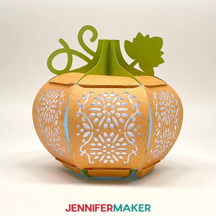 Learn how to make a 3D pumpkin lantern with JenniferMaker's tutorial! An orange paper pumpkin lantern with a green stem, vine, and leaf feature panels with intricate floral filigree designs cut out.