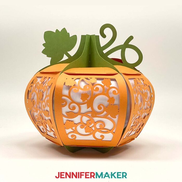 Learn how to make a 3D pumpkin lantern with JenniferMaker's tutorial! An orange paper pumpkin lantern with a green stem, vine, and leaf feature panels with intricate filigree leaf designs cut out.