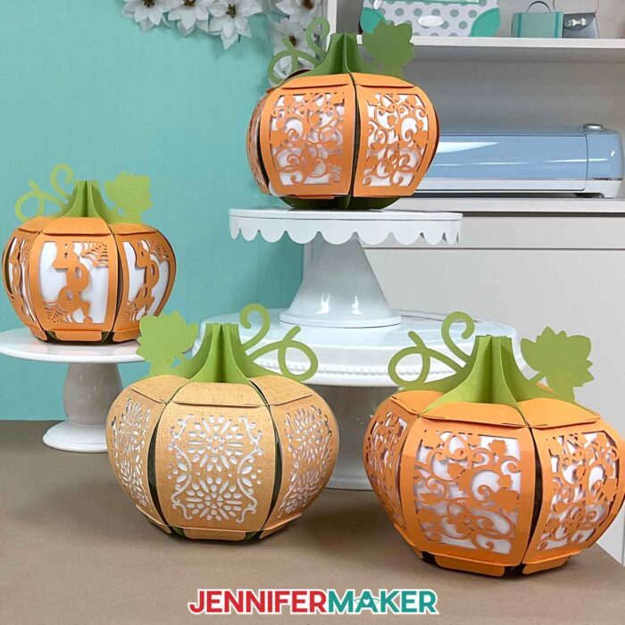 Learn how to make a 3D pumpkin lantern with JenniferMaker's tutorial! Four pumpkin lanterns, all with different designs cut from them, sit on Jennifer's craft table. Two are risen up on cake plates, and you can see her Cricut cutting machine in the background.