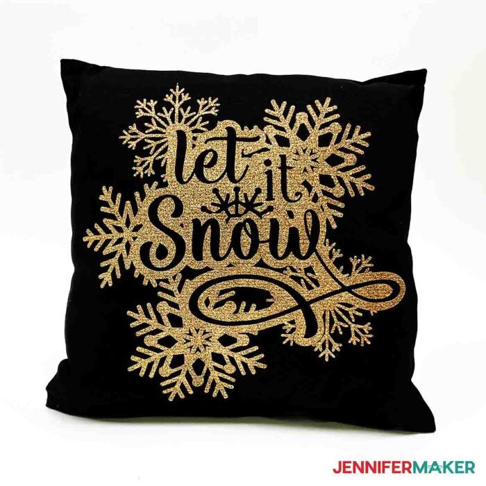 A black pillow with a gold intricate vinyl design featuring snowflakes and Let It Snow that used reverse weeding.