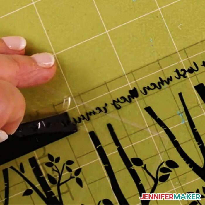 Closeup of a crafter using reverse weeding vinyl trchniques to remove excess black vinyl from small script style words that are temporarily face down on transfer tape.