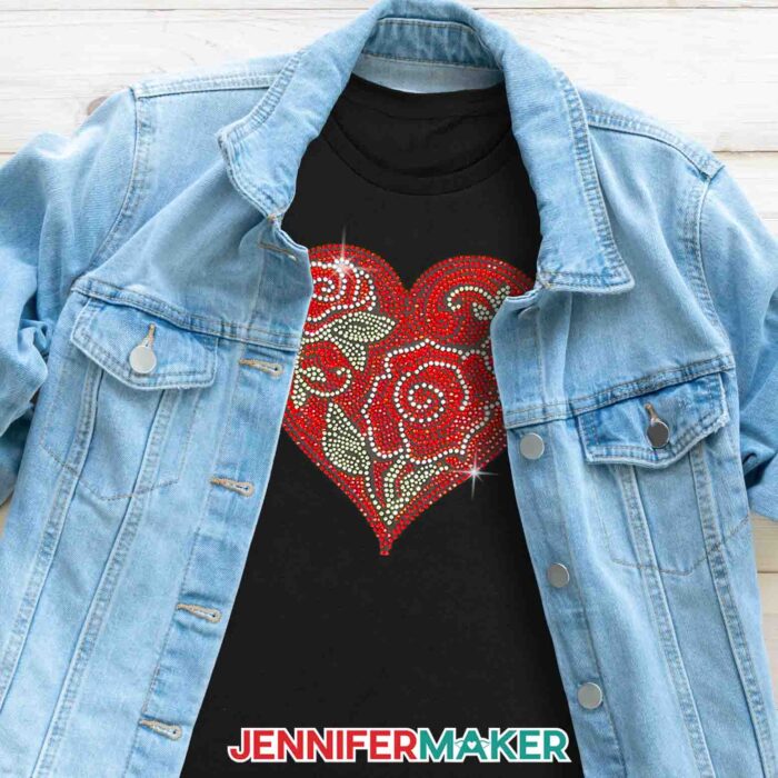 A jean jacket opens o reveal a rhinestone design of red roses and green leaves inside a red heart, sparkling on a black t-shirt. Learn to make a rhinestone template on Cricut with Jennifer Maker's tutorial!