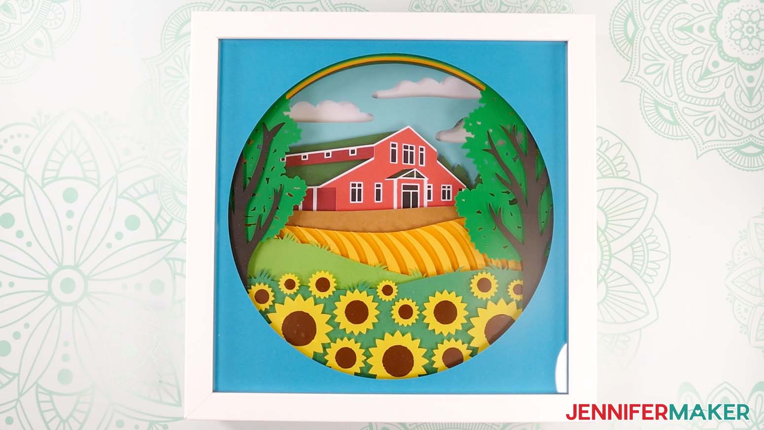 A finished summer shadow box with a barn and sunflower and tree floating pieces on a work surface