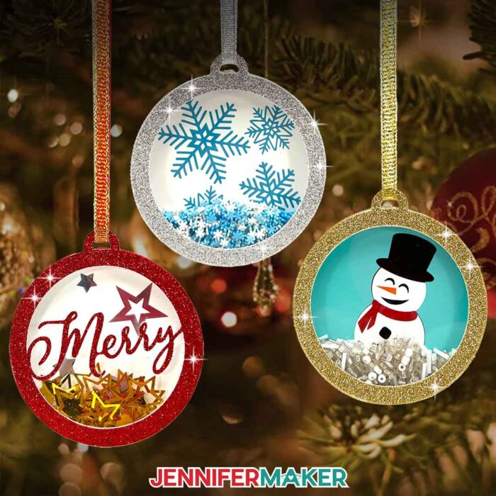 Learn how to make holiday shaker ornaments with Jennifer Maker's tutorial! Three round, glitter-framed cardstock ornaments with acetate front covers and vinyl backdrops depicting winter themes hang from a tree by ribbon. Each ornament has shiny confetti inside.
