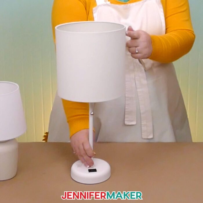 Make a DIY hidden silhouette lampshade with JenniferMaker's tutorial! Jennifer holds a small table lamp with a straight-side lampshade at her craft table.