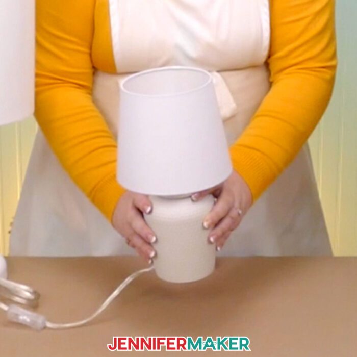 Make a DIY hidden silhouette lampshade with JenniferMaker's tutorial! Jennifer holds a small table lamp with a tapered lampshade at her craft table.