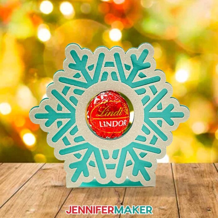 Learn how to make snowflake candy holder with JenniferMaker's new tutorial! A freestanding snowflake candy holder sits on a wooden table in front of a brightly lit Christmas tree filled with white lights and red bulb ornaments.