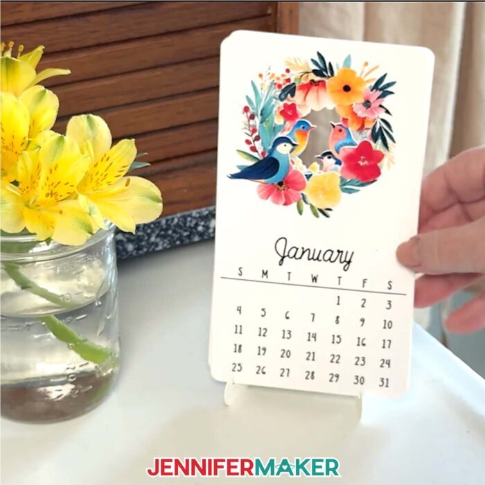 A desk calendar with birds and flowers on a cardstock stand rests on a table beside a yellow flower in a jar.