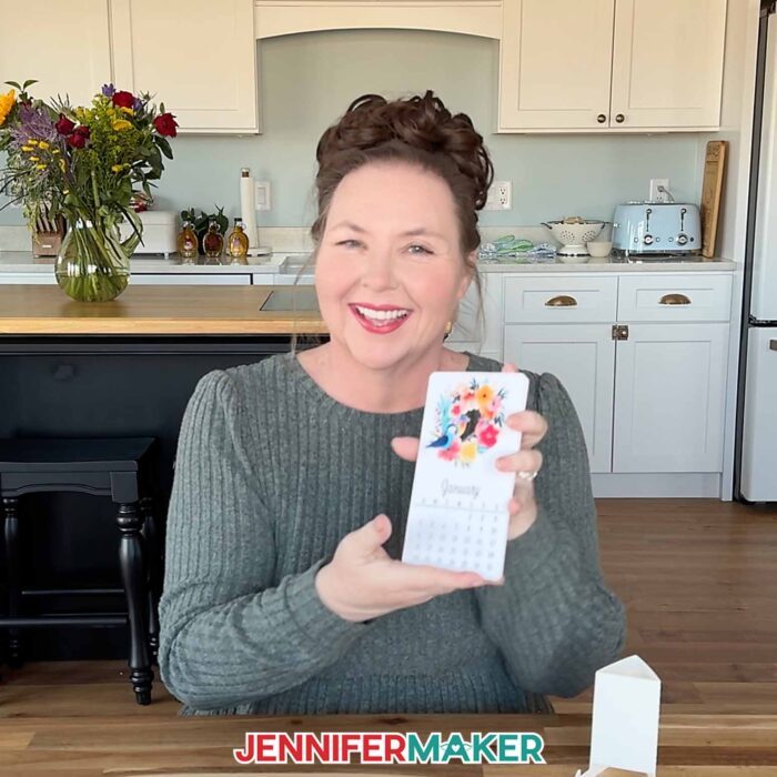 Jennifer sits at a table holding up her birds and flowers stacked desk calendar, showing the January card at the front of the stack. A cardstock stand for the calendar sits on the table.