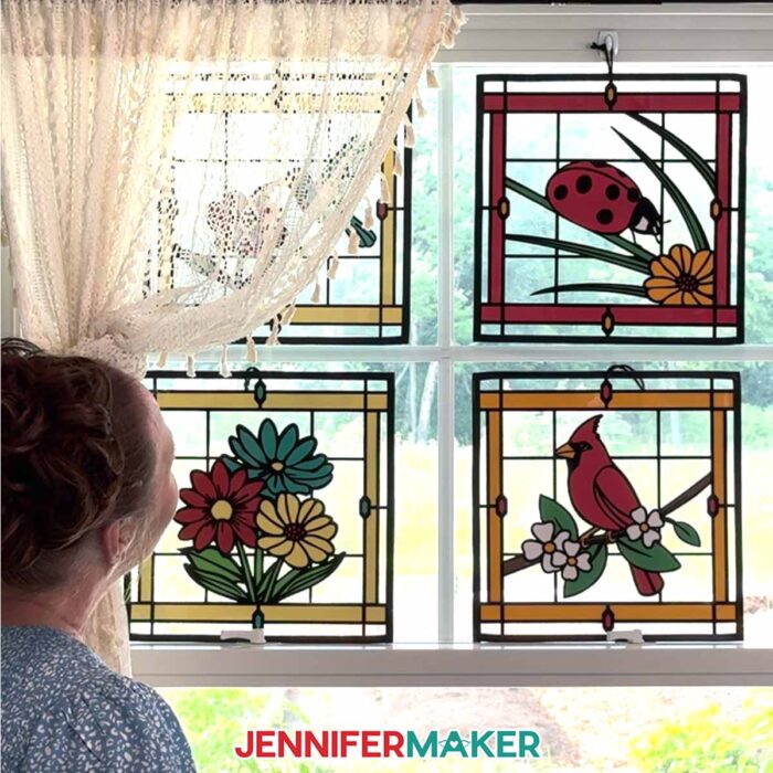 Make Stained Glass Using Paper! Jennifer Maker admires four stained glass projects hanging in a sunny window, partially obscured by a lacy beige curtain
