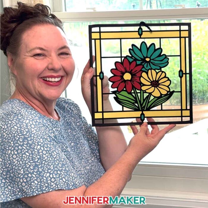 Make Stained Glass Using Paper! Jennifer Maker holds a paper, acetate, and vinyl stained glass window-inspired craft depicting a bouquet of flowers in front of a sunny window