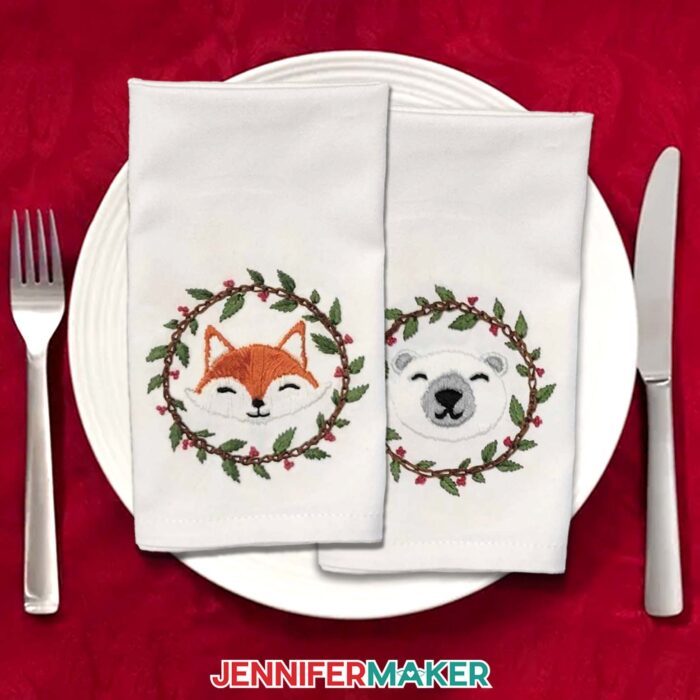 Two cloth napkins, each with Stick and Stitch Embroidery designs of a fox and a polar bear, lay on a white plate on a red holiday tablecloth