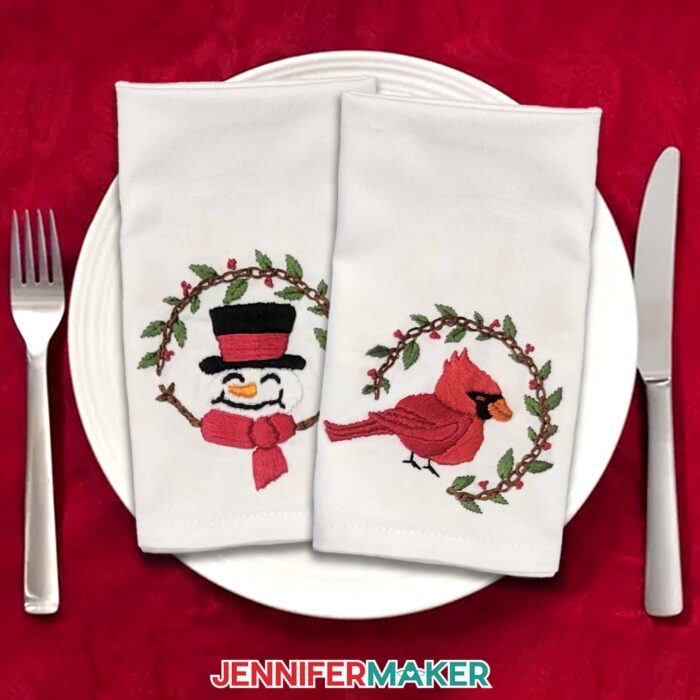 Two napkins with Stick and Stitch Embroidery sit on a white plate on a red tablecloth, one with a smiling snowman design and one with a cardinal