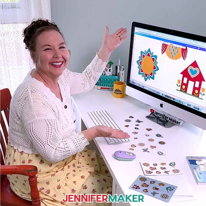 Jennifer Maker sits at her computer working on a sun mandala and red barn sticker in Canva. She has several sheets of stickers as well as some die-cut stickers spread across her desk.