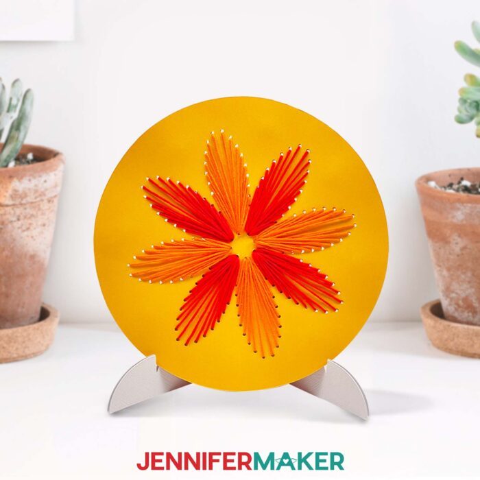 Round yellow cardstock in a stand, with a red and orange string art pattern of a flower made with a JenniferMaker tutorial.