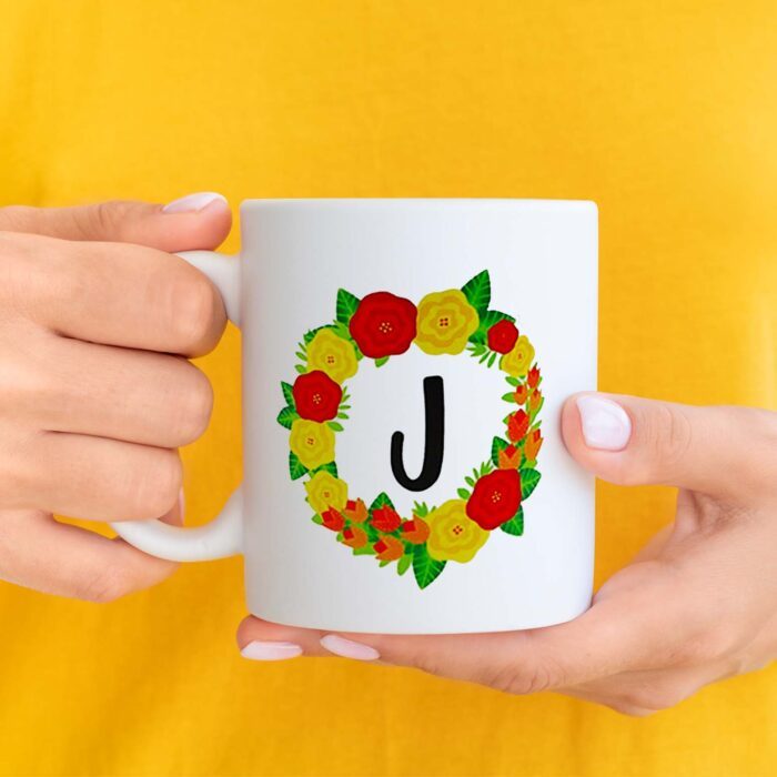 Sublimated floral mug, held against a background of yellow. A wreath design encircles a bold "J" and is made up of red, orange, and yellow flowers. Learn how to sublimate mugs with JenniferMaker's tutorial!