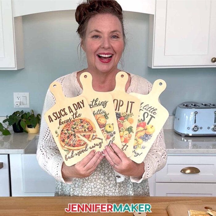 Jennifer Maker holds up four sublimated charcuterie boards with food-themed designs and captions while standing in her kitchen.