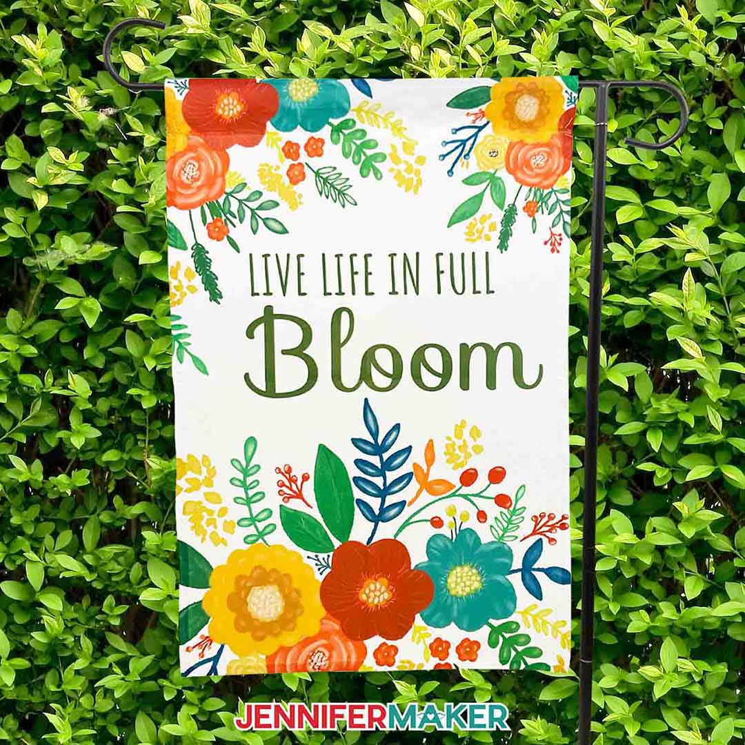 Brightly colored sublimated garden flag hanging on a black hanger against a backdrop of bright green foliage. The flag reads "live life in full bloom". Learn to make your own sublimation garden flag with Jennifer Maker's new tutorial!