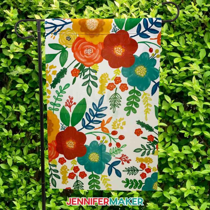A beautifully colored floral garden flag hangs on a black hanger against a wall of thick green leaves. Make your own sublimation garden flag with JenniferMaker's tutorial! 