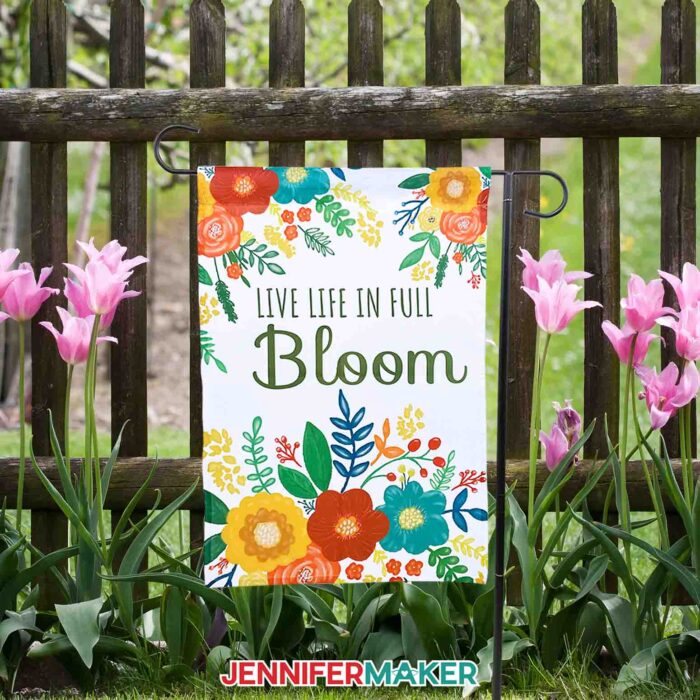 Make your own sublimation garden flag with JenniferMaker's tutorial! A brightly colored garden flag with flowers and the words "Live Life in Full Bloom" hang next to a wooden garden fence, surrounded by pink flowers. 