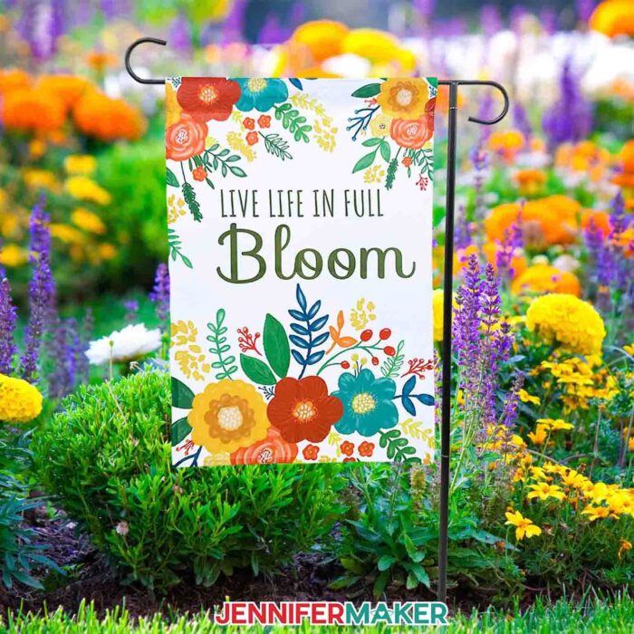 A brightly colored floral garden flag hangs in a garden full of spring flowers. The flag reads "Live Life in Full Bloom". Make your own sublimation garden flag with JenniferMaker's tutorial! 