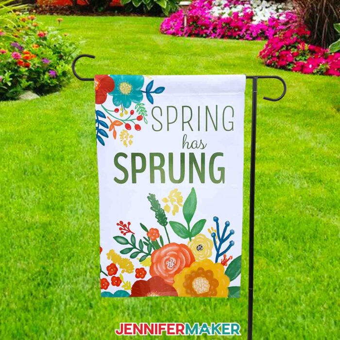 A sublimated garden flag reading "Spring Has Sprung" hangs against a backdrop of lush green lawn and springtime flowers. Make your own sublimation garden flag with JenniferMaker's tutorial! 