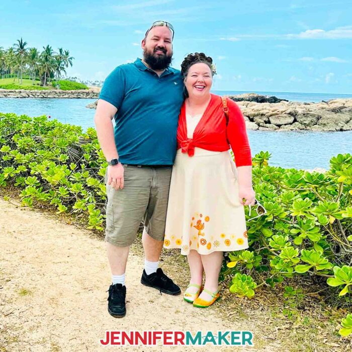 Greg Reese and Jennifer Maker standing together with big smiles in front of a greenery and water scene in Hawaii. Jennifer is wearing a light yellow dress she sublimated designs on to.