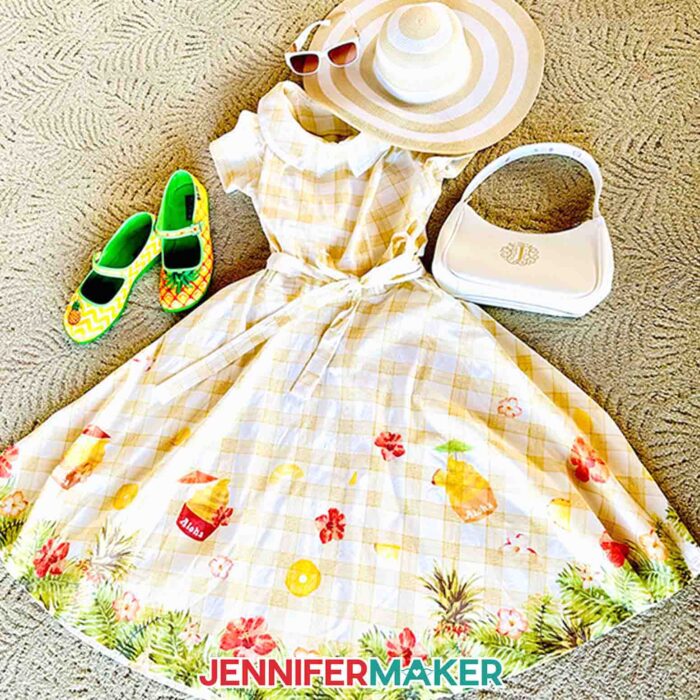 A flat laid white vintage style dress with yellow checked pattern and Dole Whip-inspired designs sublimated on the skirt, with a monogrammed white purse, pineapple shoes, white sunglasses, and beach hat.
