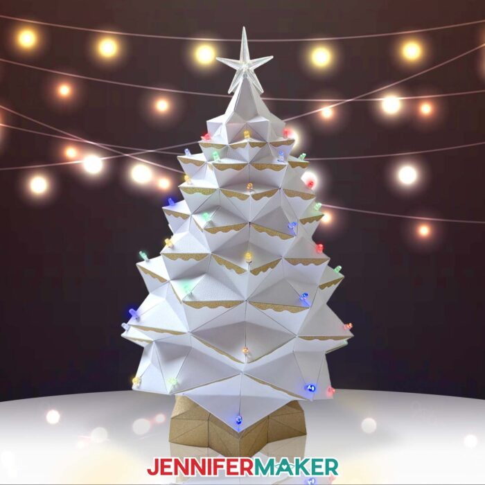 Learn how to make a light-up tabletop Christmas tree for the holidays with JenniferMaker's tutorial! An elaborate white cardstock Christmas tree glows with multicolored lights against a dark backdrop studded in twinkle lights.