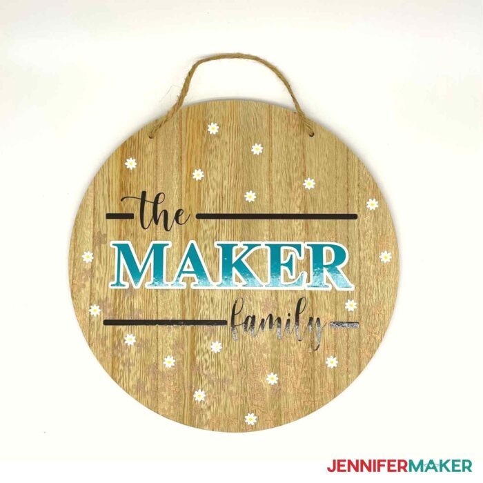 Round wooden sign from Target Dollar Spot DIYs with the Maker family decal in blue, black, and white decal and vinyl flowers.