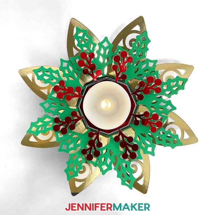 Intricately cut cardstock Tea Light Holder inspired by a holiday poinsettia, made from green, red, and gold foil cardstock and filled with a glowing LED tea light