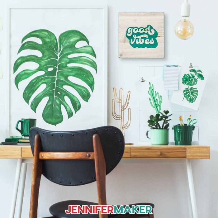 A wooden plaque with "good vibes" in green hangs in a cute plant-inspired workspace with other artwork of monstera leaves and cacti, green desk accessories, and lots of bright light. Learn how to use transfer tape with vinyl projects with JenniferMaker's new tutorial!