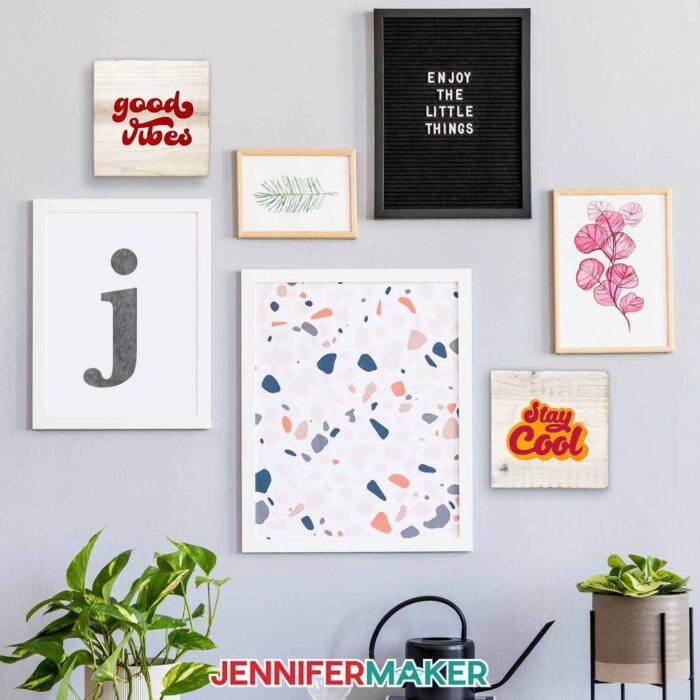 Two wooden plaques with "good vibes" in red and "stay cool" in red and yellow hang on a gray wall with other artwork of botanical and abstract subjects, as well as one that says "enjoy the little things" and one with a giant "J". Plants line the bottom of the photo. Learn how to use transfer tape with vinyl projects with JenniferMaker's new tutorial!