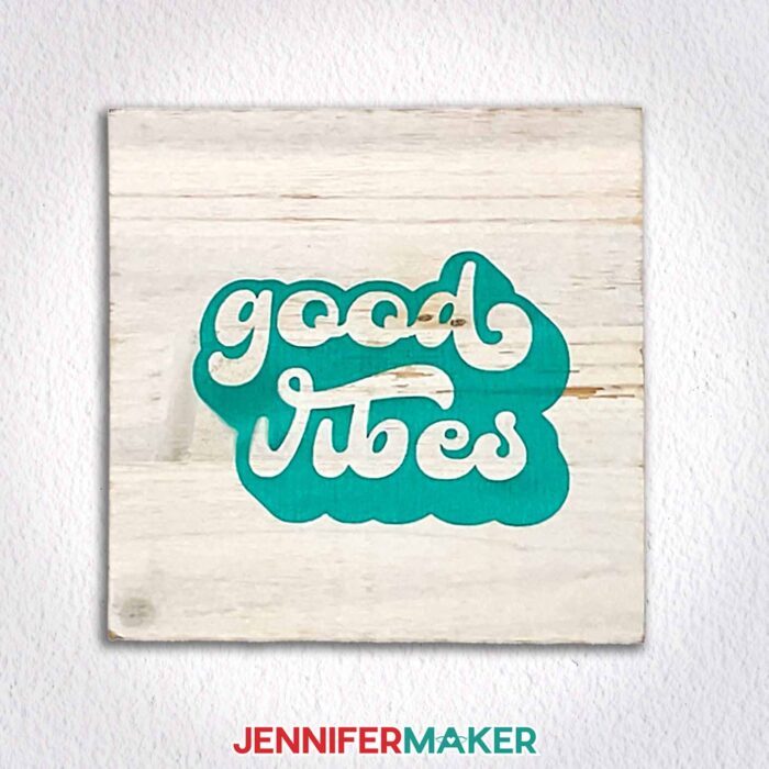 A wooden plaque with "good vibes" outline in green vinyl hangs on a white wall. Learn how to use transfer tape with vinyl projects with JenniferMaker's new tutorial!