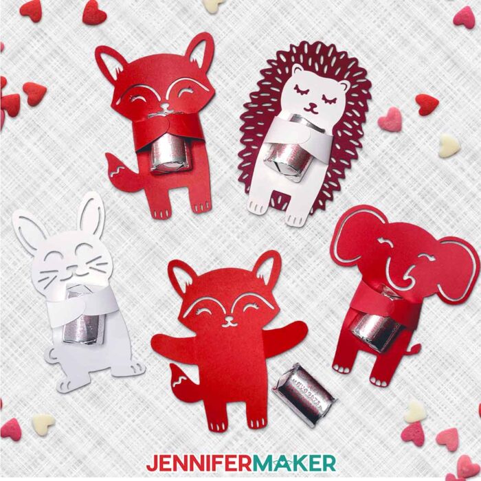 A collection of cute paper animal Valentine Candy Huggers made of red and white cardstock sits on a white background, each holding a wrapped piece of chocolate candy.